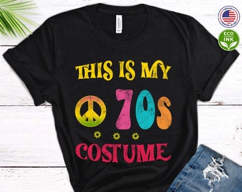 Puede incluir: Camiseta negra con el texto "This is my 70s Costume" en letras de colores con un signo de paz y flores. La camiseta está sobre una superficie de madera blanca con un par de zapatillas blancas y unos vaqueros azules.