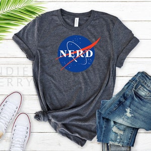 May include: A dark heather gray t-shirt with a blue and red NASA-inspired logo that says "NERD" in white text.