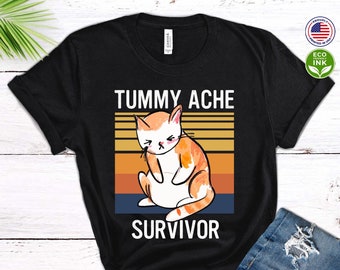 May include: Black t-shirt with a vintage-style graphic of a cat with a tummy ache. The text "Tummy Ache Survivor" is printed below the cat.