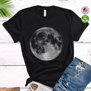 May include: Black t-shirt with a large, detailed image of the moon in shades of gray. The shirt is laid flat on a white wooden surface with a pair of white sneakers and blue jeans.