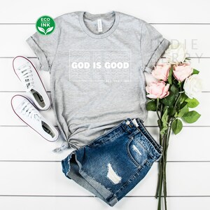 May include: A gray t-shirt with the text "GOD IS GOOD" in a white rectangular design. The phrase "All the time" is below the main text. The shirt is paired with denim shorts and white sneakers. A bouquet of pink and white roses is also present.