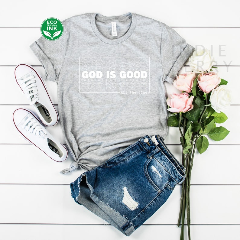 May include: A gray t-shirt with the text "GOD IS GOOD" in a white rectangular design. The phrase "All the time" is below the main text. The shirt is paired with denim shorts and white sneakers. A bouquet of pink and white roses is also present.