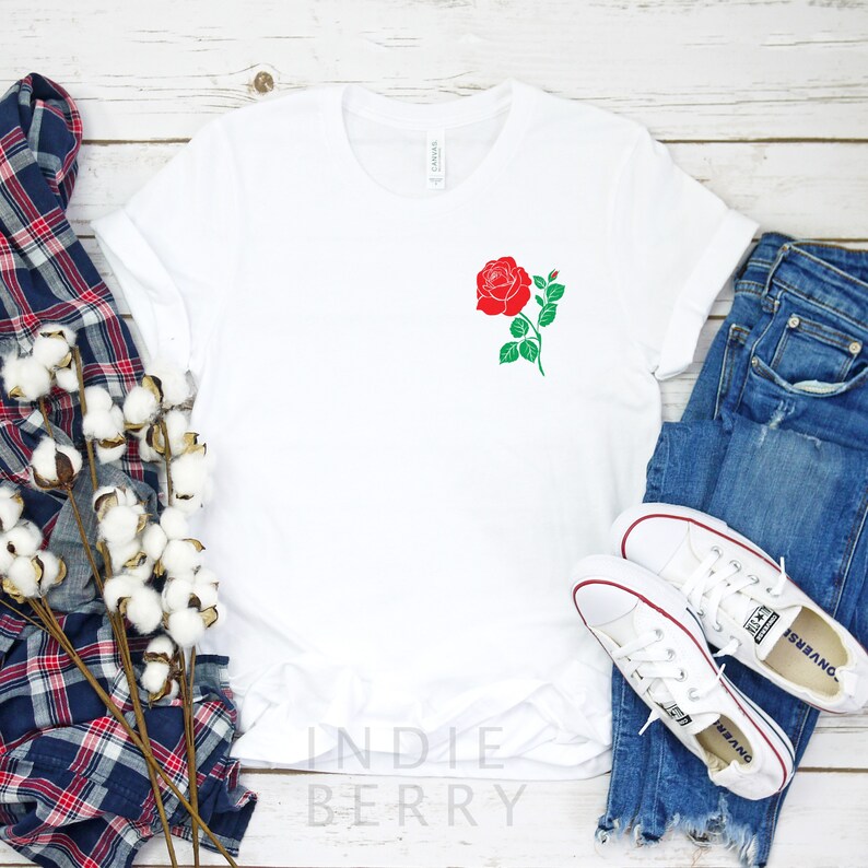 May include: White t-shirt with a red rose graphic on the left chest. The shirt is laying flat on a white wooden surface with a blue and red plaid shirt and a pair of white sneakers.