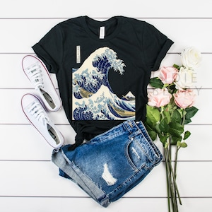 May include: Black t-shirt featuring a print of Hokusai's "The Great Wave off Kanagawa" artwork. The shirt is paired with denim shorts and white sneakers. A bouquet of pink roses adds a touch of color to the scene. The shirt has a classic crew neck.