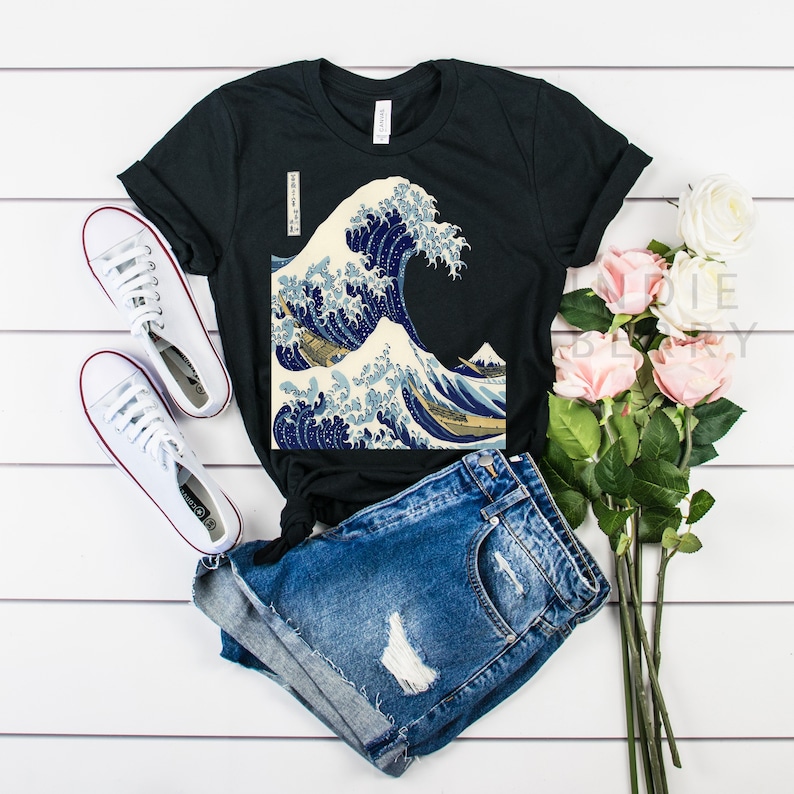 May include: Black t-shirt featuring a print of Hokusai's "The Great Wave off Kanagawa" artwork. The shirt is paired with denim shorts and white sneakers. A bouquet of pink roses adds a touch of color to the scene. The shirt has a classic crew neck.
