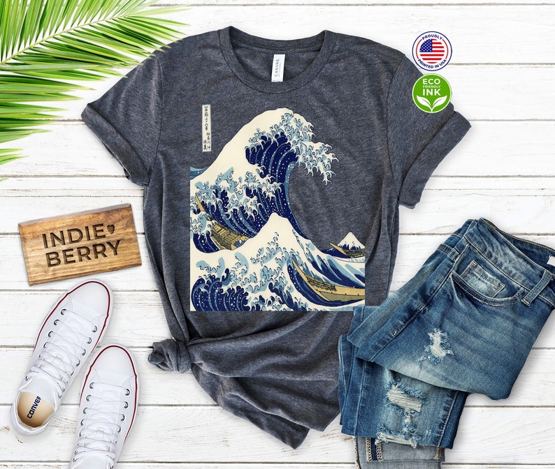 May include: Dark gray t-shirt featuring a print of Hokusai's "The Great Wave off Kanagawa." The design showcases a large wave with blue and white hues, with a mountain in the background. The shirt is paired with blue jeans and white sneakers.