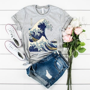 May include: A gray t-shirt featuring a print of Hokusai's "The Great Wave off Kanagawa" artwork. The shirt is paired with denim shorts and white sneakers. A bouquet of pink and white roses adds a touch of color to the scene.