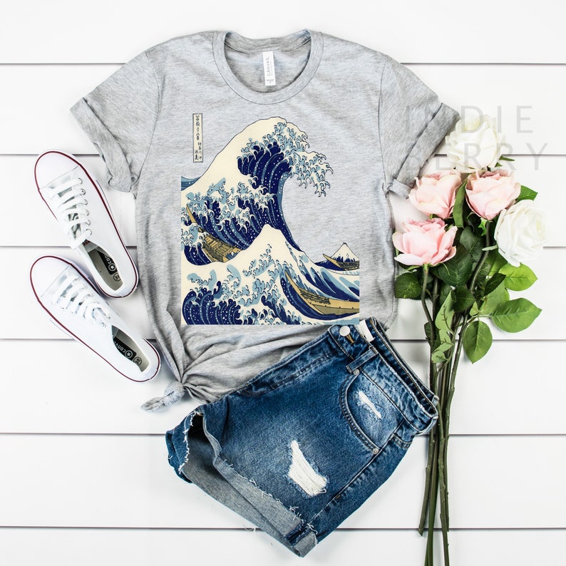 May include: A gray t-shirt featuring a print of Hokusai's "The Great Wave off Kanagawa" artwork. The shirt is paired with denim shorts and white sneakers. A bouquet of pink and white roses adds a touch of color to the scene.