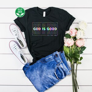 May include: Black t-shirt with a rainbow graphic that says "God is Good - All the time". The shirt is laid out on a white wooden surface with a pair of white sneakers, blue jeans, and pink flowers.