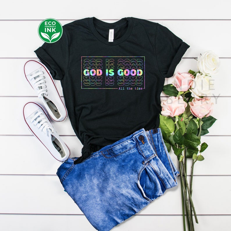 May include: Black t-shirt with a rainbow graphic that says "God is Good - All the time". The shirt is laid out on a white wooden surface with a pair of white sneakers, blue jeans, and pink flowers.