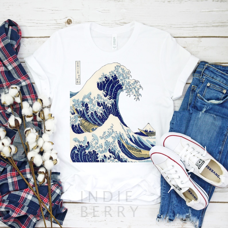 May include: White t-shirt featuring a classic Japanese wave print in shades of blue and cream. The design includes a mountain and boats. The shirt is paired with blue jeans and white sneakers.
