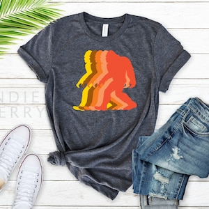May include: A dark heather grey t-shirt with a retro-style graphic of a Bigfoot walking. The graphic is in shades of orange, yellow, and brown.