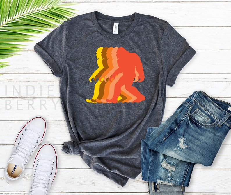 May include: A dark heather grey t-shirt with a retro-style graphic of a Bigfoot walking. The graphic is in shades of orange, yellow, and brown.