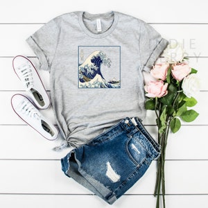 Op de afbeelding: Grijs T-shirt met een grafische print van de Grote Golf voor Kanagawa, een beroemde Japanse houtsnede van Hokusai. De print toont een grote golf met de berg Fuji op de achtergrond.