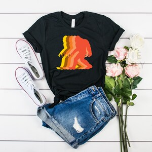 May include: A black t-shirt with a retro-style graphic of a Bigfoot walking. The graphic is in shades of orange, yellow, and brown. The t-shirt is laid out on a white wooden surface with a pair of white sneakers and a bouquet of pink roses.