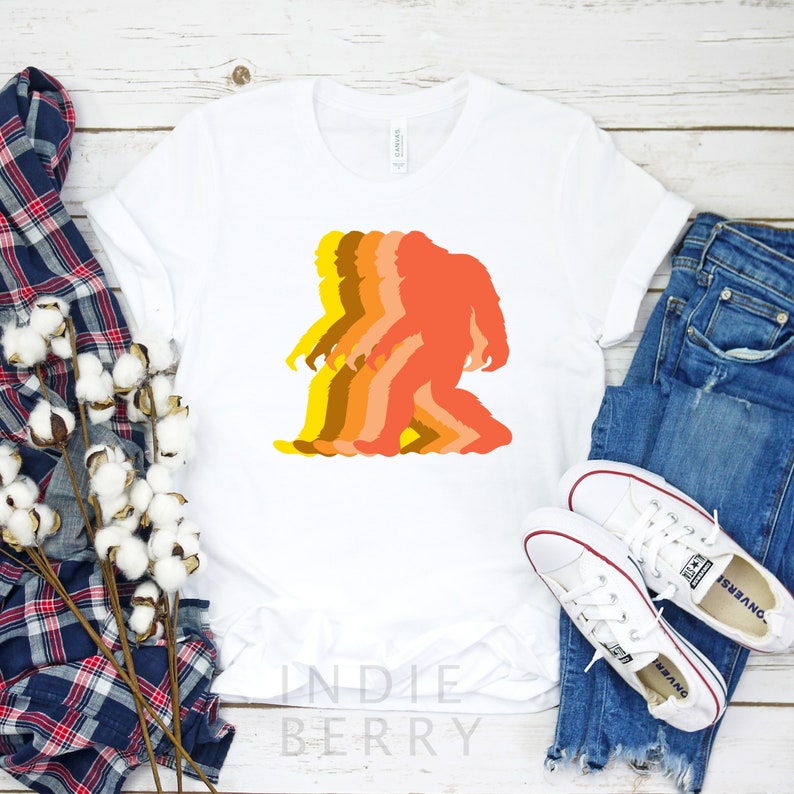 May include: White t-shirt with a graphic of a silhouette of a Bigfoot walking. The silhouette is made up of five different shades of orange, yellow, and brown. The text "Indie Berry" is printed on the bottom of the shirt.