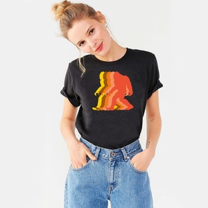 May include: A black t-shirt with a retro-style graphic of a Bigfoot walking. The graphic is in shades of orange, yellow, and red.