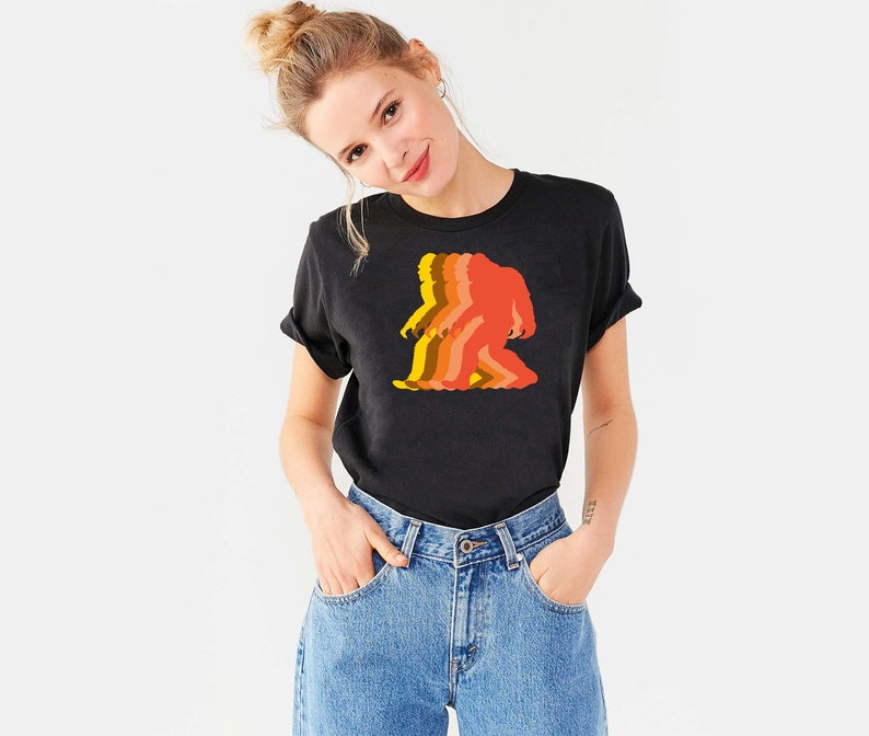 May include: A black t-shirt with a retro-style graphic of a Bigfoot walking. The graphic is in shades of orange, yellow, and red.