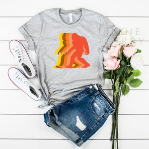 May include: A gray t-shirt with a retro-style silhouette of a Bigfoot in shades of orange, yellow, and brown.