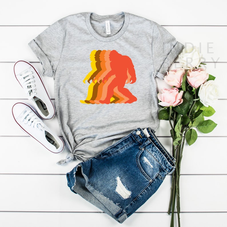 May include: A gray t-shirt with a retro-style silhouette of a Bigfoot in shades of orange, yellow, and brown.