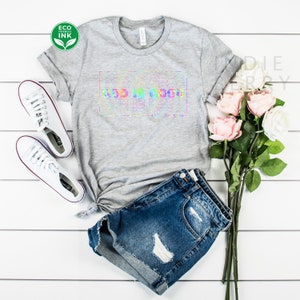 May include: A gray t-shirt with a rainbow tie-dye design that says "God is good" in a white box. The shirt is styled with denim shorts and white sneakers.