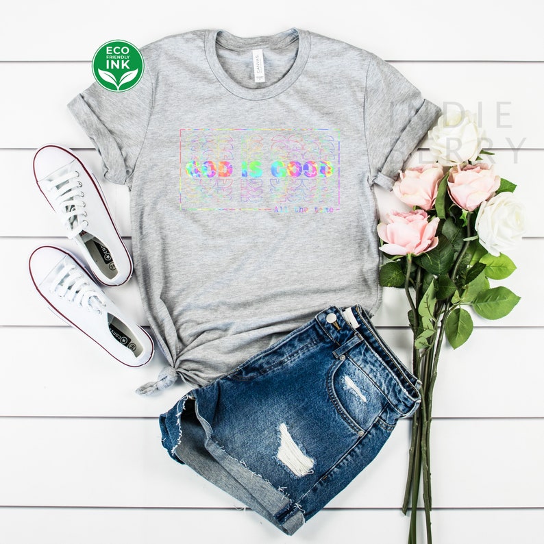 May include: A gray t-shirt with a rainbow tie-dye design that says "God is good" in a white box. The shirt is styled with denim shorts and white sneakers.