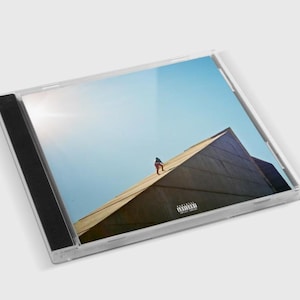 May include: A CD case featuring album art with a person standing on a building's roof under a bright blue sky. The album art has a minimalist design with a person in red and black clothing. The case is transparent.