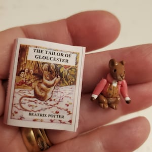 May include: A small, brown mouse figurine wearing a pink jacket with gold buttons. The figurine is holding a small book titled "The Tailor of Gloucester" by Beatrix Potter.
