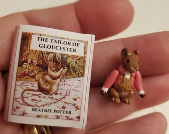 Könnte beinhalten: Eine kleine, braune Mausfigur, die eine rosa Jacke mit goldenen Knöpfen trägt. Die Figur hält ein kleines Buch mit dem Titel "Der Schneider von Gloucester" von Beatrix Potter.