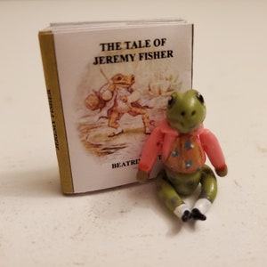 May include: A miniature figurine of a frog wearing a pink jacket and orange vest, sitting next to a small book titled "The Tale of Jeremy Fisher." The book cover features an illustration of a frog. The figurine has green skin and white socks.
