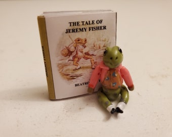 Könnte beinhalten: Eine Miniaturfigur einer Froschfigur mit rosa Jacke und orangefarbener Weste, neben einem kleinen Buch mit dem Titel "The Tale of Jeremy Fisher". Das Buchcover zeigt eine Froschillustration. Die Figur hat grüne Haut und weiße Socken.