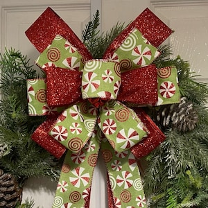 May include: A festive Christmas wreath featuring a large bow with red glitter and green ribbon. The green ribbon is patterned with white and red peppermint candies. Pinecones and evergreen branches complete the holiday decoration.
