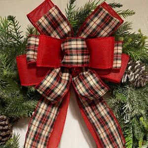 May include: A Christmas wreath with a prominent red bow, patterned with a plaid design in red, black, and beige. The bow is set against a backdrop of green pine needles and pine cones, creating a festive holiday decoration.