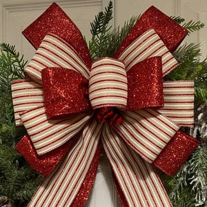 May include: A festive Christmas wreath bow featuring a large, multi-layered bow with red glitter and striped ribbon. The bow is set against a backdrop of green pine needles and a white door, creating a holiday theme.