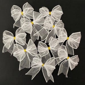 May include: A group of ten white sheer bows with gold centres. The bows are arranged in a cluster on a black background.