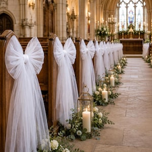 May include: A row of wooden church pews decorated with large white tulle bows tied to the ends. Each pew features a sheer white fabric bow, while gold metal lanterns containing white pillar candles and greenery are placed along the stone aisle.