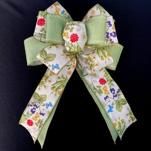 May include: A decorative bow crafted from two distinct ribbons. One ribbon is a solid light green, while the other features a white background with a colourful botanical print of red, yellow, and purple flowers, green leaves, and small blue butterflies.