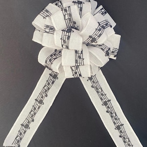 May include: A decorative white bow with black musical notes printed on the fabric. The bow has multiple loops and two long streamers. The musical notes are evenly spaced along the ribbon, creating a musical theme.
