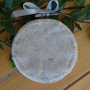 May include: A circular, pale grey felt decoration with white stitching around the perimeter. It is hung by a silver-tone metal ring and a grey ribbon. The initials "M." are embroidered on the felt. Decorative item.