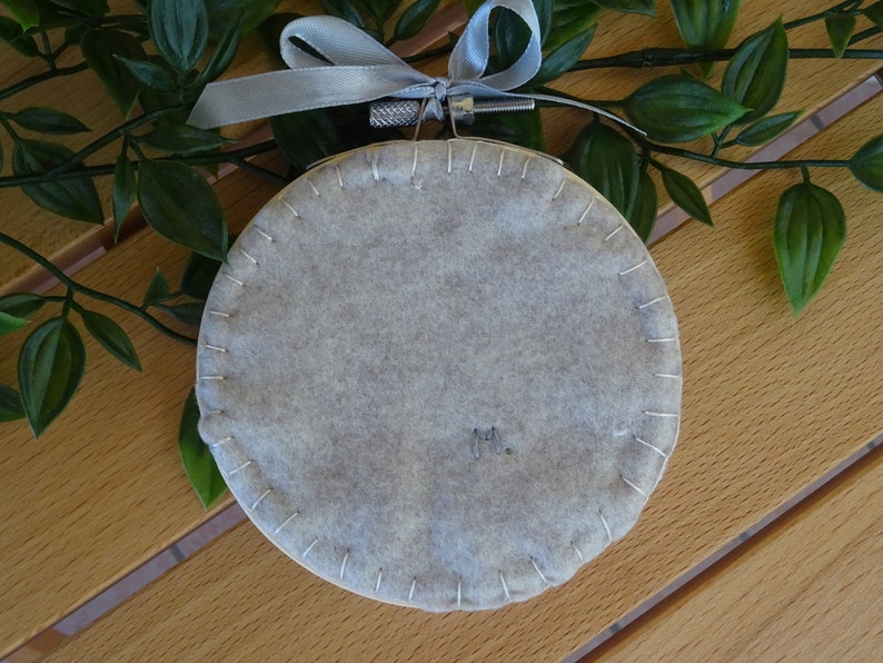 May include: A circular, pale grey felt decoration with white stitching around the perimeter. It is hung by a silver-tone metal ring and a grey ribbon. The initials "M." are embroidered on the felt. Decorative item.
