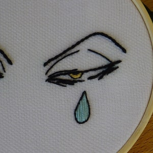 May include: Embroidered artwork featuring stylized eyes with black thread and yellow accents. A single teal teardrop is stitched below the eye. The design is set in a wooden embroidery hoop, showcasing a minimalist, emotional theme.