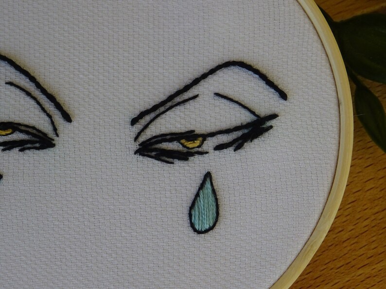 May include: Embroidered artwork featuring stylized eyes with black thread and yellow accents. A single teal teardrop is stitched below the eye. The design is set in a wooden embroidery hoop, showcasing a minimalist, emotional theme.