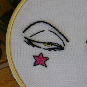 May include: Embroidered face on white fabric, set in a wooden embroidery hoop. The face features black lines for the eyebrows and eyelashes, a yellow eye, and a pink star. A craft item.