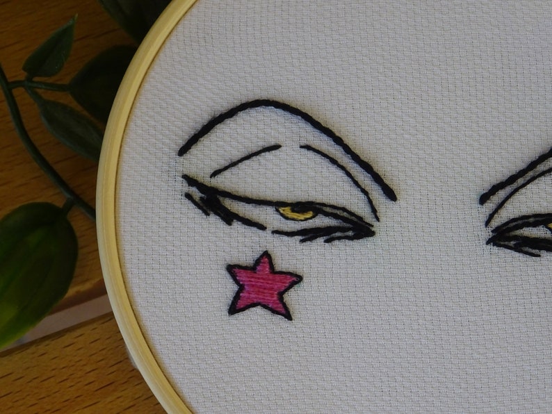 May include: Embroidered face on white fabric, set in a wooden embroidery hoop. The face features black lines for the eyebrows and eyelashes, a yellow eye, and a pink star. A craft item.