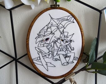 May include: Black and white embroidery hoop featuring a cartoon image of two characters holding an umbrella. The characters are a demon with horns and a cat wearing a top hat. The embroidery is on white fabric.