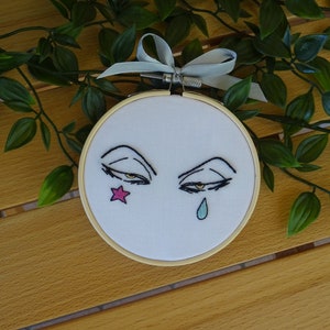 May include: Embroidered artwork in a wooden hoop features stylized eyes with a star and teardrop. The eyes are outlined in black with yellow irises. A pink star is on one side, and a blue teardrop on the other. A grey ribbon is attached.