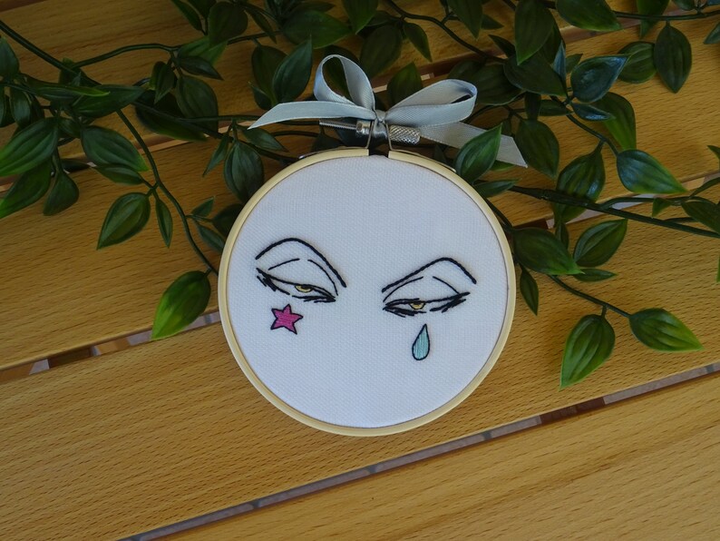 May include: Embroidered artwork in a wooden hoop features stylized eyes with a star and teardrop. The eyes are outlined in black with yellow irises. A pink star is on one side, and a blue teardrop on the other. A grey ribbon is attached.