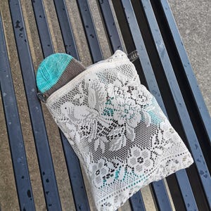 May include: A white lace zippered pouch with a teal and brown knitted hat partially visible. The pouch features a floral design and a black zip. The hat has a teal crown and a brown brim.
