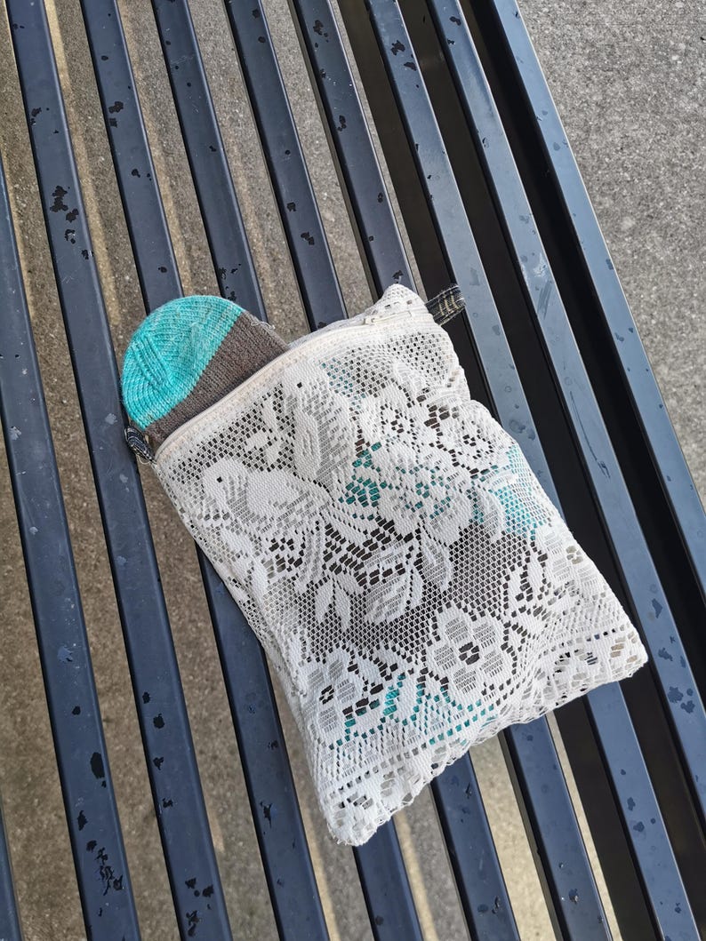 May include: A white lace zippered pouch with a teal and brown knitted hat partially visible. The pouch features a floral design and a black zip. The hat has a teal crown and a brown brim.