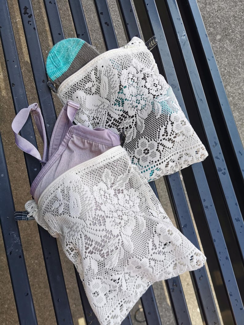 May include: Two open, white lace bags on a dark bench. One bag contains a teal and grey sock, the other a lavender bra. The bags have a floral lace pattern and are partially unzipped, revealing the contents.